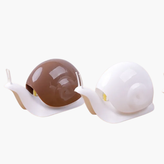 Trois porte savon en céramique en forme d’escargot sur une surface en bois avec un fond de rideau blanc.