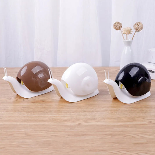 Trois porte savon en céramique en forme d’escargot sur une surface en bois avec un fond de rideau blanc.